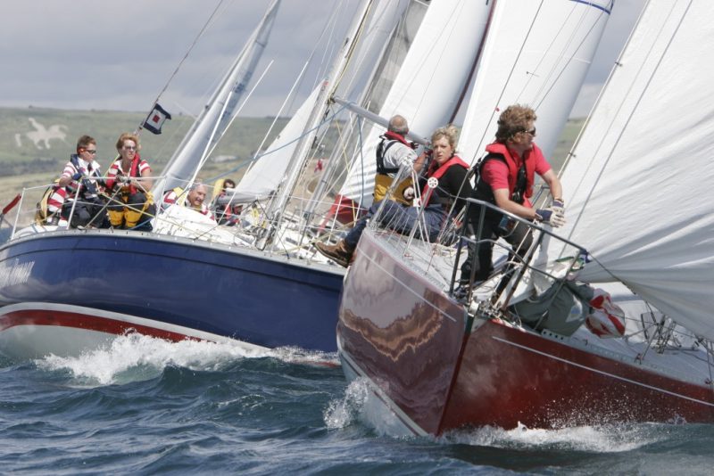 two sailing yachts racing on the sea with several people on the deck of each one and the weymouth white horse visible on the hillside behind