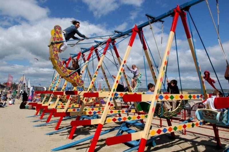 Wooden swingboats on a sandy beach painted in cream and red with multicoloured diamonds, there is a woman and a young girl in a yellow boat swinging up to the left