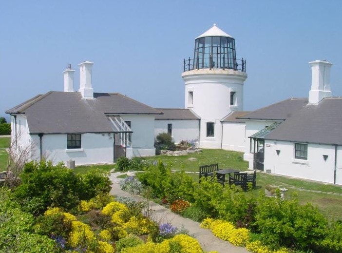 small white lighthouse in between two one-storeywhitewashed cottages with grey tiled roods, with a small garden infront filled with flowers 