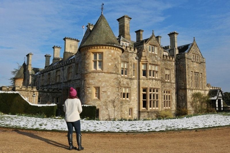 Emily wearing a white wooly jumper, pink wool hat, black skinny jeans and knee-high black Hunter wellington boots standing in front of an historic manor house built from beige stone with a circular turret on the corner and grey tiled roof. the ground  and wall in front of the house is covered with snow. 