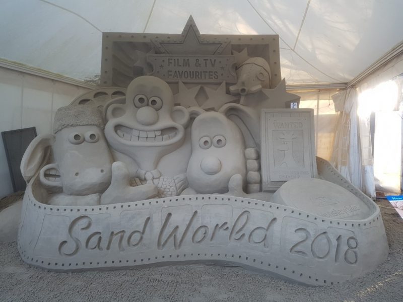 large sand sculpture with the faces of the cartoon characters Wallace, Gromit and Shawn the Sheep carved into it and a banner along the bottom which reads: Sandworld 2018