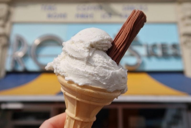 hand holding an ice cream cone with a single scoop of plain white ice cream with a chocolate flake in front of an out of focus ice cream parlour facade