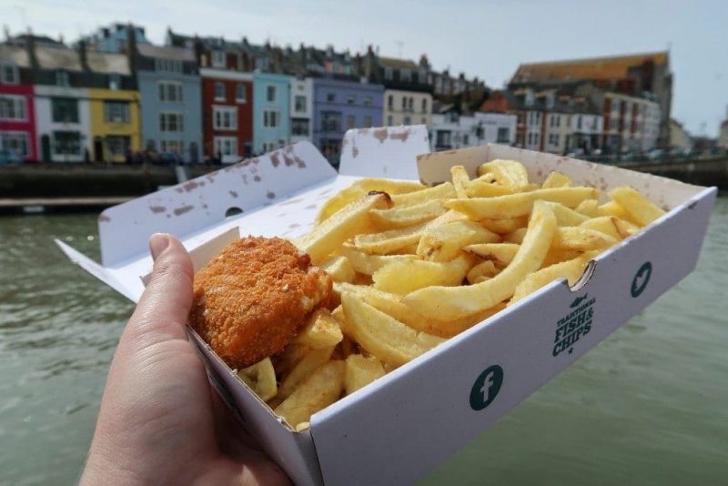 hand holding a white paper box filled with thick cut chips and a small breaded fishcake in front of a habrour with a row of narrow terraced houses painted in different colours, taken on a grey cloudy day.