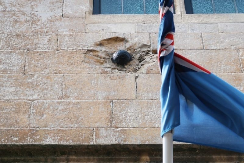 blue flag in front of a grey stone wall where there is a metal canonball lodged into a deep dent