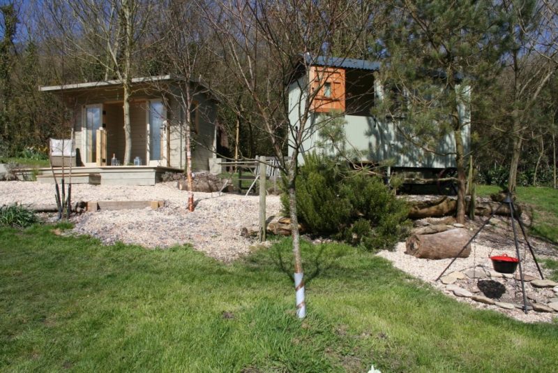 metal shepherds hut painted light green behind some trees next to a small wooden hut with a gravel garden area in front of both and a small firepit with cooking apparatus 