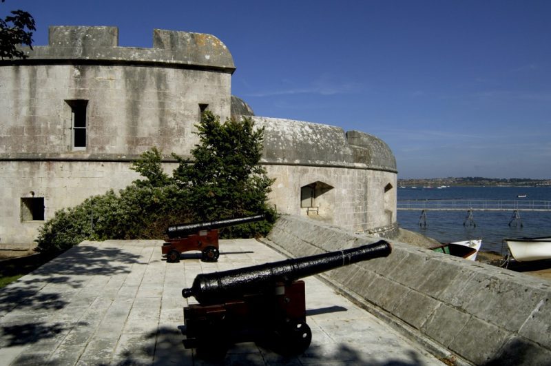 ramparts of a grey stone castle with a large round turret beyond and two black canons lined up on the edge of the ramparts