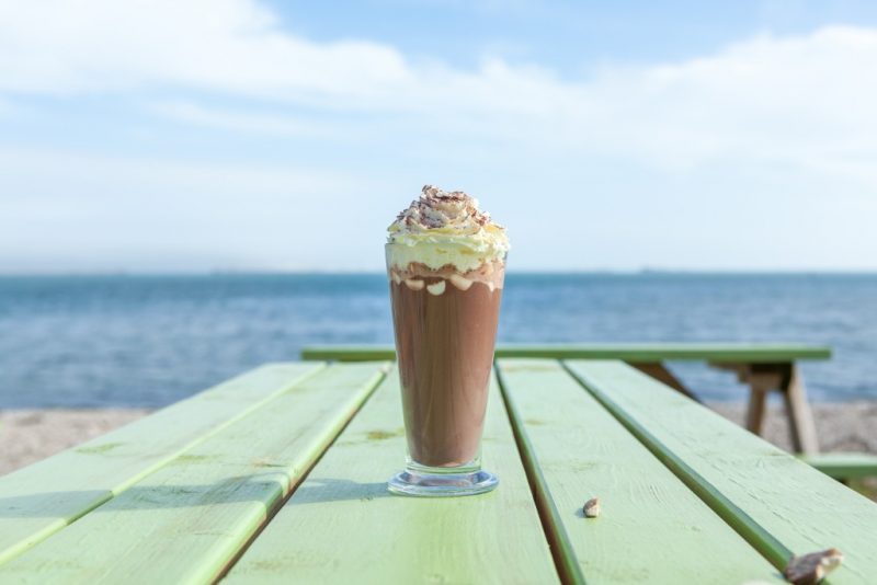 chocolate milkshake in a glass topped with whipped cream on a green wooden table outside next to a shingle beach with a view of the blue sea beyond on a sunny day