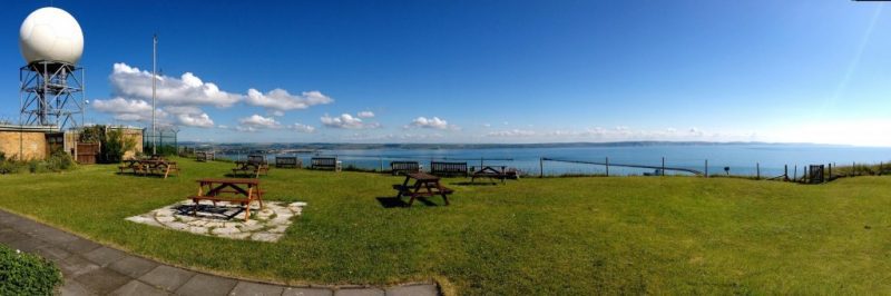 large grassy lawn with several wooden picnic benches dotted around on a cliff top with a view of the blue sea on a sunny day