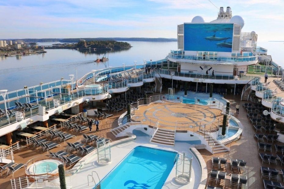 Outdoor top deck of the Regal Princess cruise ship with an empty blue rectangular swimming pool in the centre and a raised circular stage area beyond that with a big screen showing a film with dolphins swimming underwater above that. there is ocean around the cruise ship with a few islands in Stockholm Harbour on a sunny day with blue sky.