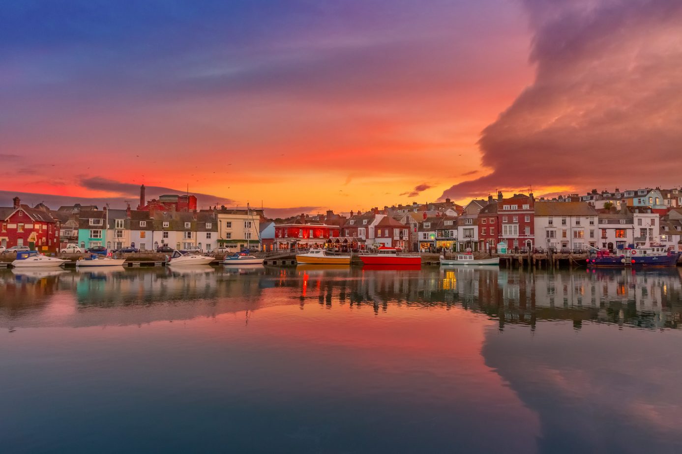 Looking across Weymouth Harbour at sunset with a row of colourful fishing boats moored along the far bank in front of a long row of terraced narrow houses all painted in different colours with the sky orange and purple overhead.  50 Unique Things to do in Weymouth