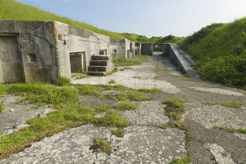 Verne High Angle Battery on Portland in Dorset, partly ruined concrete buildings and rooms with steps arranged around a wide paved area with grassy verges on either side