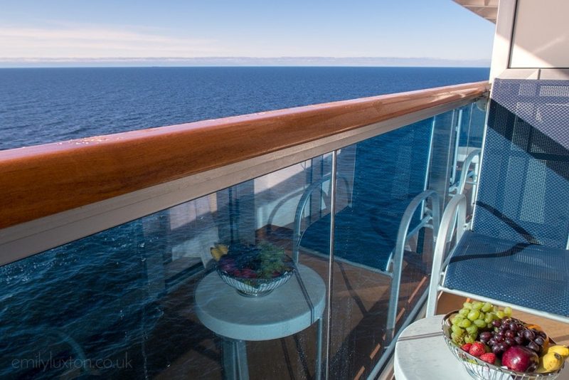 Regal Princess balcony with view of the sea and a silver bowl of fruit on a small white circular table on the balcony next to the glass barrier with wooden edging - cabin reviews