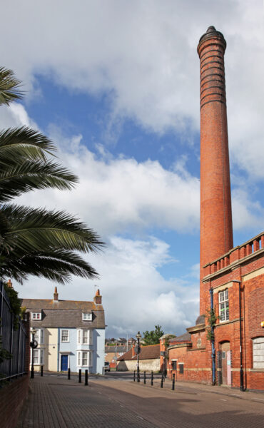 Red brick building with a very tall chimney tower overlooking smaller stone cottages