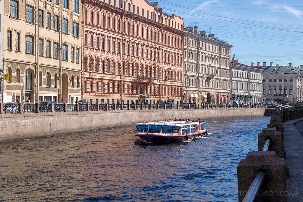 river in St Petersburg with a small ferry boat on it and tall yellow and pink apartment buildings along the far bank