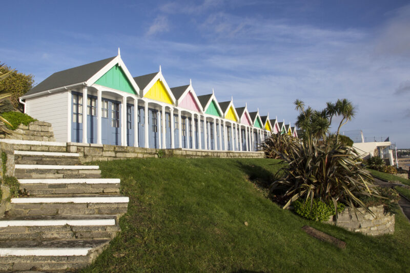 White wooden beach huts with different coloured pastel roofs and a garden in front