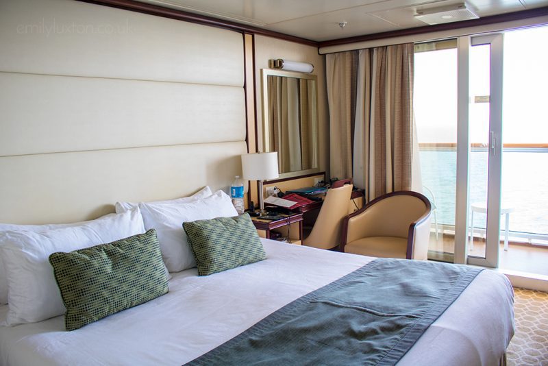 Interior of a cruise ship cabin with a double bed with white bedspread and light green cushions and throw. the walls are cream and there is a large french door with a view of the sea beyond. Regal Princess balcony cabin