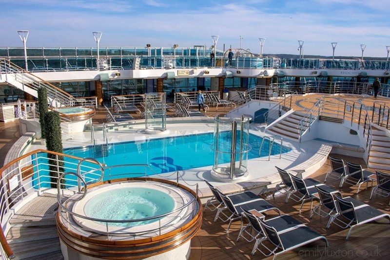 Top outside deck of the Regal Princess cruise ship with wooden floor covered with metal and blue canvas sunbeds. there is a rectangular blue swimming pool in the centre of the deck with the Priness Cruises logo of an abstract hair with long wavy hair painted on the floor of the pool. in front of the camera there is a white circular hot tub with bright blue water. the deck and pool are empty on an early morning with blue sky above. 