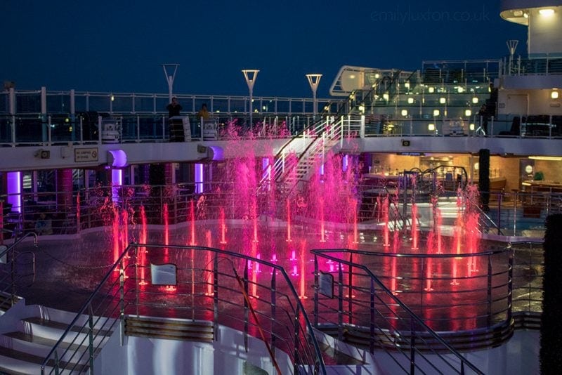 deck of a cruise ship outdoors at night with a large fountain with multiple jets of water all lit in red and pink. Regal Princess review