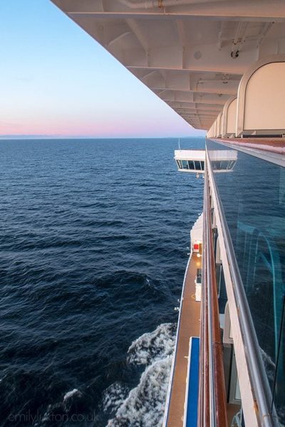 Looking along the side of a cruise ship from one of the cabins, there is a long strip of glass along the centre of the ship with a wooden deck below and empy blue sea to the left with a clear blue sky with pink on the horizon just after sunset. 