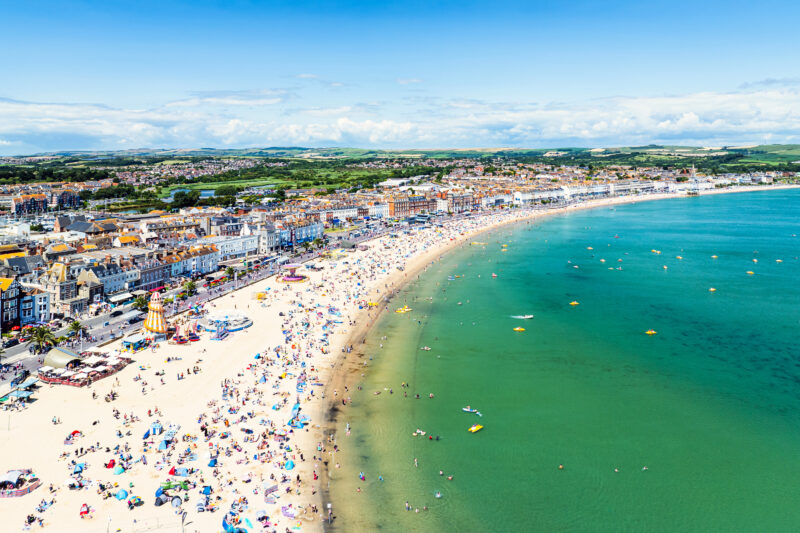 Aerial view of a long sandy beach crowded with people with very turquiose water and Weymouth town behind
