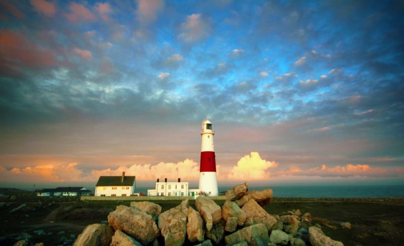 tall white lighthouse with a red stripe around the middle on portland bill in weymouth with rocks in the foregound and the sea just visible behind taken at sunset with pink clouds in the sky