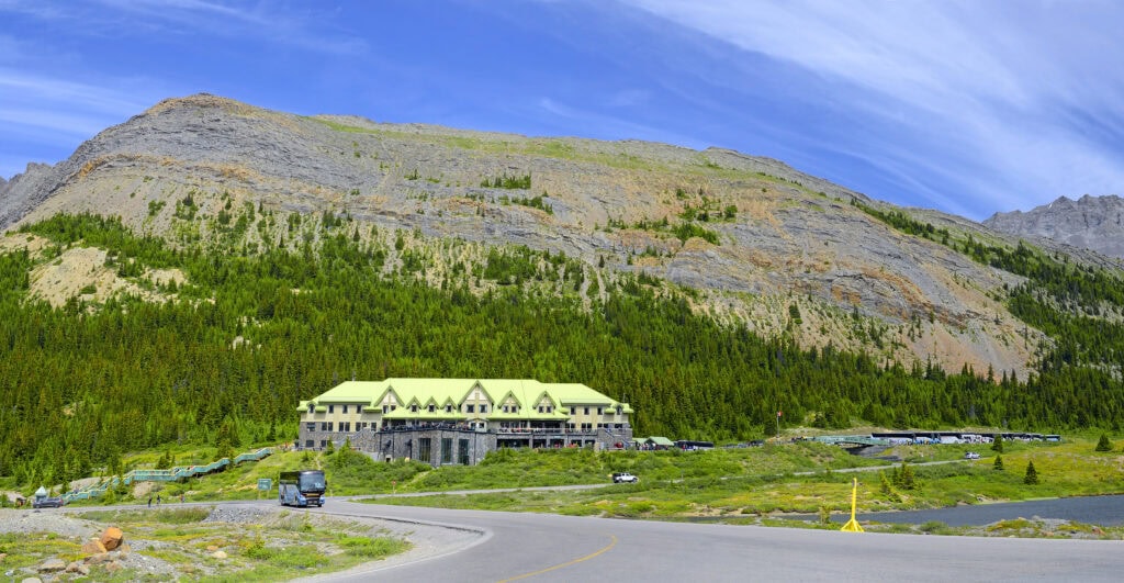 Large grey stone building with a green roof infront of a rocky mountain in summer