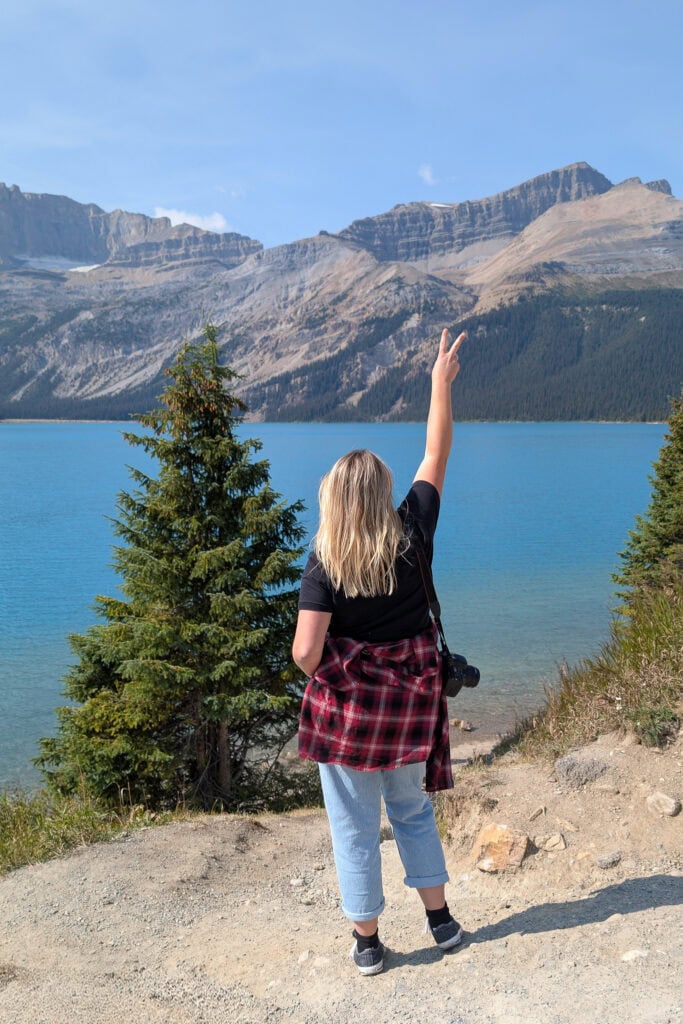 Woman with blonde hair standing beside a fir tree in front of a bright blue lake with mountains on the far side. She is wearing jeans and a black t-shirt with a red plaid shirt tied around her waist.