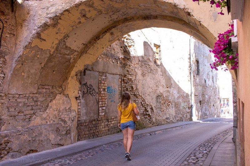 emily walking in a cobbled street under an archway in vilnius wearign a yellow t shirt and denim shorts - Simple summer city break packing list.