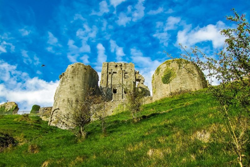 looking up at the ruins of a grey stone castle on top of a grassy hill on a very sunny day 