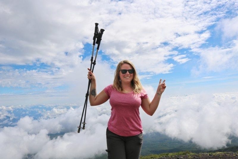 emily wearing a pink sports top and sunglasses with her blonde hair down around her shoulders standing on a mountainside with clouds behind her on a sunny day. she is holding two walking poles in one hand and making a peace sign with the other. 