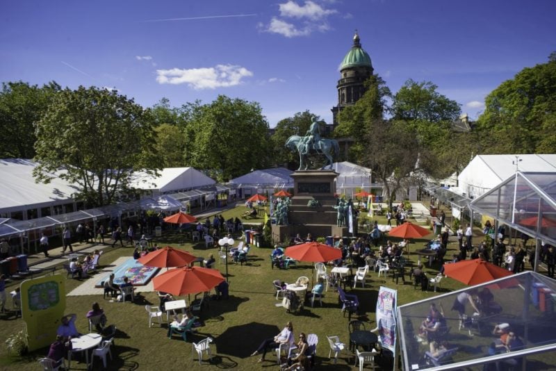 aerial shot of a lawn with many large white marquees around the sides and several outdoor tables with orange umbrellas. There is a statue of a man on a horse in the centre of the park. 