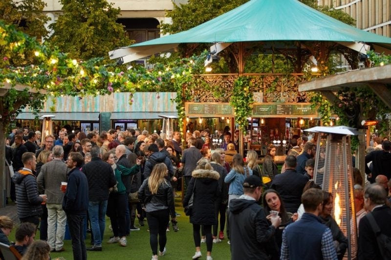 courtyard area with fake grass on the floor surrounded bu wooden pop up stalls and bars, there is a large crowd of people on the grass holding drinks. Edinburgh Fringe Tips