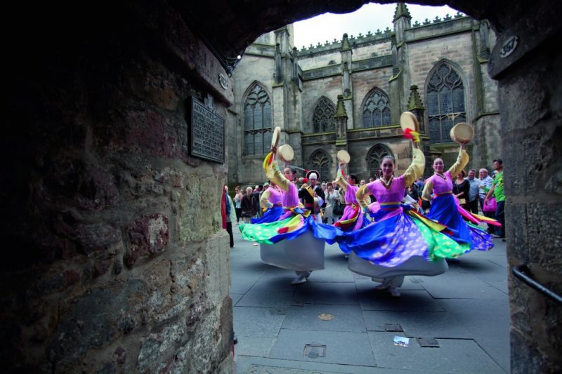 Group of female dancers spinning around so that the blue and green skirts of their dresses flare out and waving tambourines in the air in front of a church in Edinburgh at the Fringe Festival - viewed through a stone tunnel in the Old Town. 