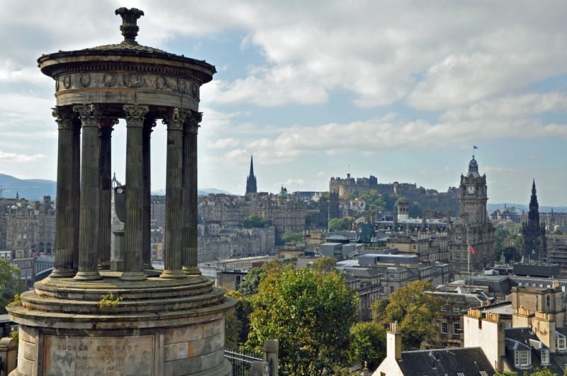 Edinburgh skyline viewed from Calton Hill on a warm sunny day with blue sky above. In the foreground is a small, circular stone monument with a domed top - the Dugald Stewart monument. Tips for your first Edinburgh Festival