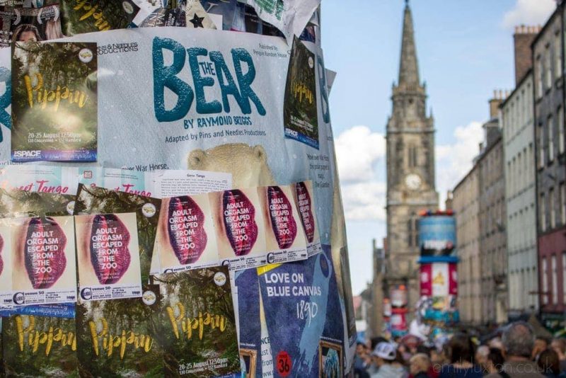 Pillar covered in many different posters and flyers for shows at the Edinburgh Fringe festival with a crowded street out of focus behind