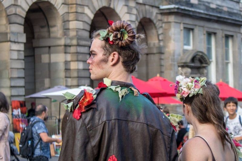 two street performers seen from beind, the man is wearing a brown leather jacket covered with flowers and leaves and has curled goat horns on his head, the woman has flowers in her long brown hair.