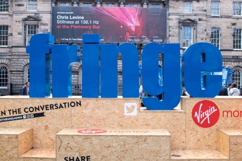 large sign with blue letters reading "Fringe" resting on wooden boxes in the street with a grey stone building behind
