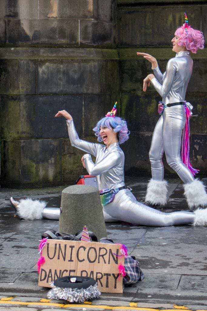 two street performers in edinburgh wearing shiny siver bodysuits with pink ribbon tails and unicorn horns. One has a pink wig and the other has a purple wig and is doing the splits. A cardboard sign in front reads: Unicorn Dance Party