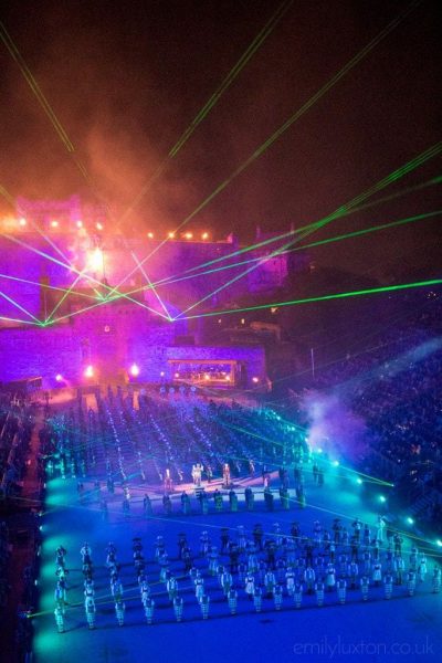 Night scene with a castle lit in bright pink with green laser-style lights pointing up from it. there is a large crowd in front lit in green and blue. Edinburgh Military Tattoo