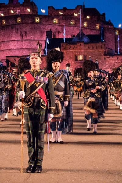 Military band wearing traditional uniforms and standing in formation in front of a stone castle lit up in purple at night. Edinburgh Military Tattoo