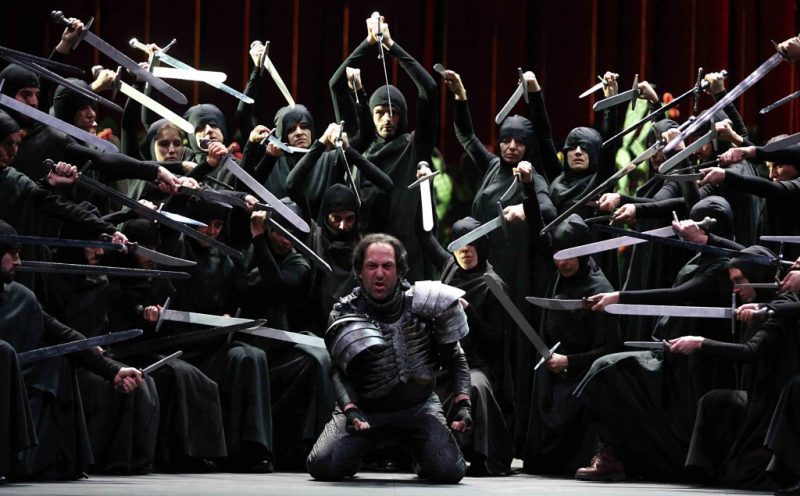 darkly lit stage with a man wearing armour kneeling in front surrounded by a laerge group of people wearing black and pointing swords at the man. 