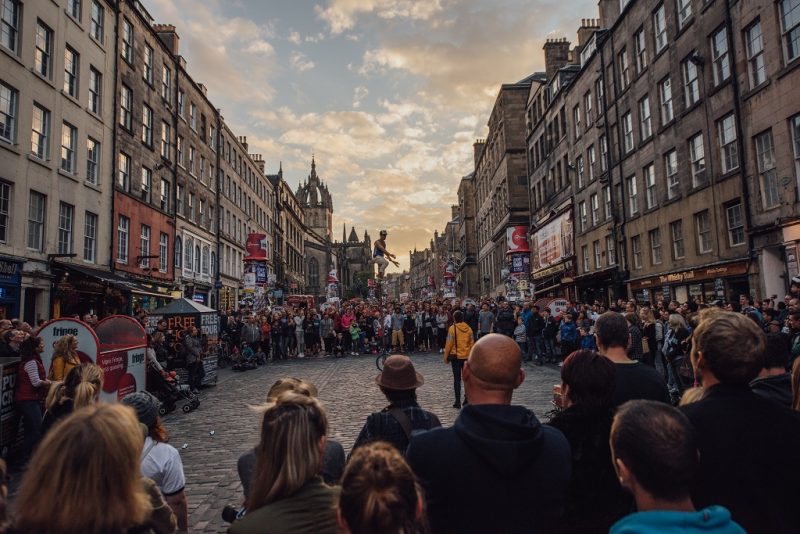 large crowd of people in a circle on a cobbled street between shops watching a man ride a unicycle