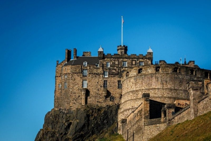 exterior of Edinburgh castle, a brown stone building with a circular section in front and a small turret in the centre on top of a rocky hill, taken from below on a sunny day with blue sky