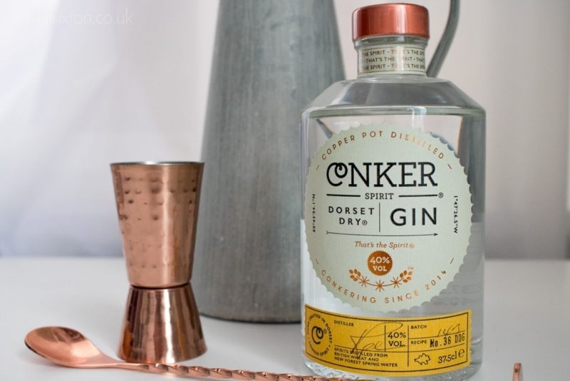 Close up of a glass bottle of Conker Spirit Dorset Dry Gin on a white table top next to a brushed metal vase and a copper coloured shot measurer and long cocktail spoon. 