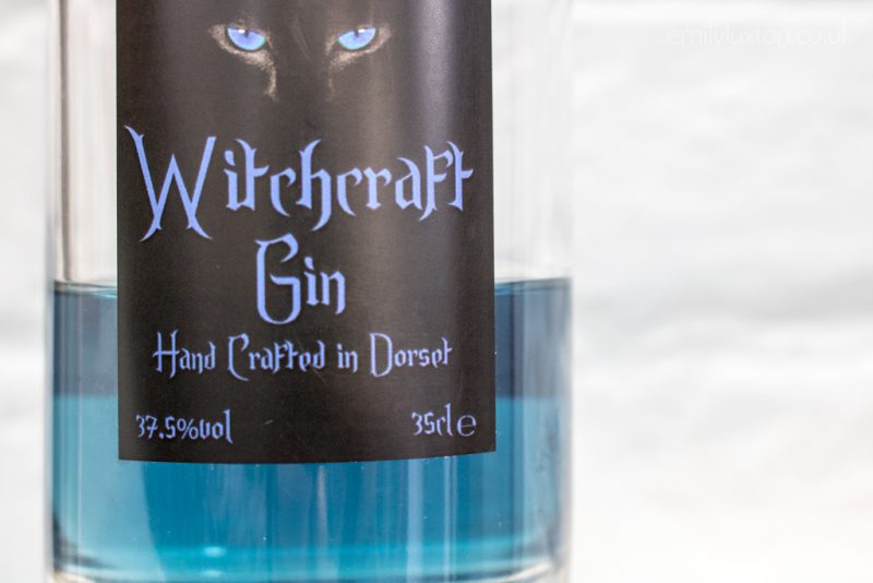 Close up of a glass bottle half filled with bright blue liquid. there is a black label with the words Witchcraft Gin Handcrafted in Dorset written on it in purple letters. 