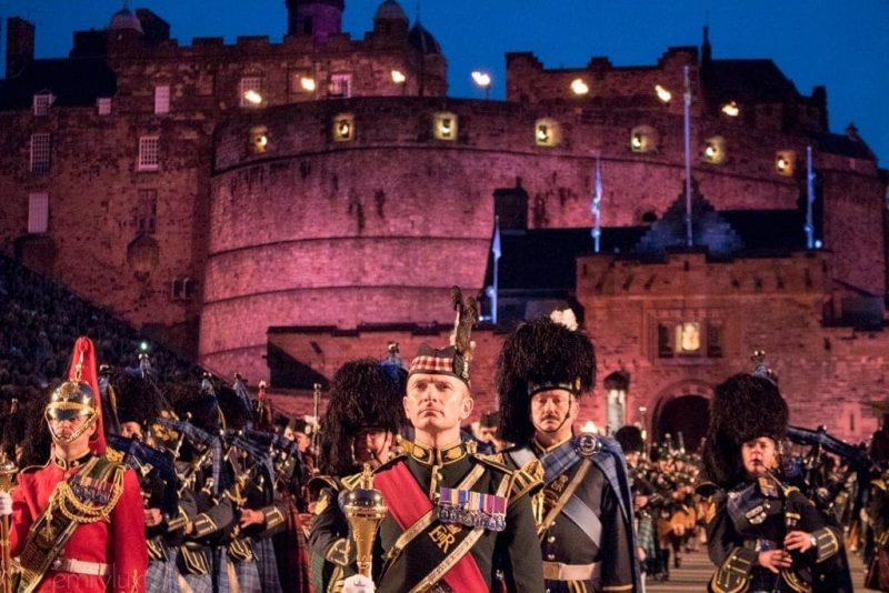 Close up of a military marching band at the Edinburgh Military Tattoo. The band are wearing the Scottish militery dress and kilts and are stood in front of the historic, rounded stone castle which is lit with purple light,  