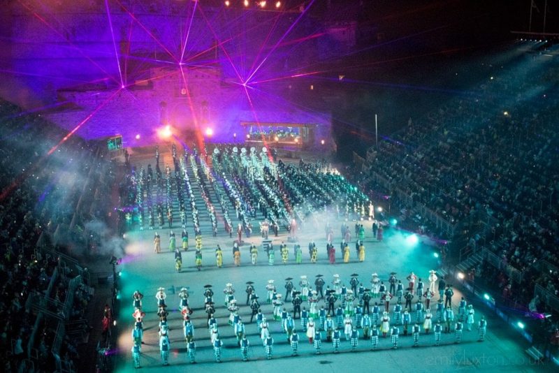 Aerial view of a huge military band standing in front of an old stone castle lit with purple and pink strobe lighting with stands of spectators on either side at the Edinburgh Military Tattoo