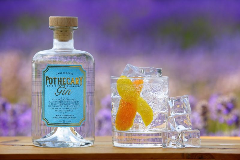 Close up of a glass bottle of Pothecary Gin with a blue and gold label next to a short tumbler glass filled with gin, tonic, ice, and a twist of orange peel, stood on a wooden table top with a field of purple lavendar out of focus behind.