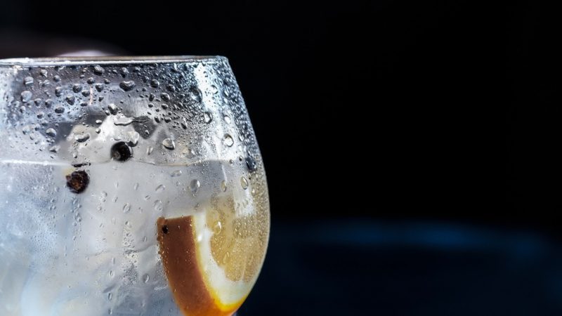 Close up of a large balloon glass of gin and tonic with ice, peppercorns and a slice of lemon against a black background. best dorset gin