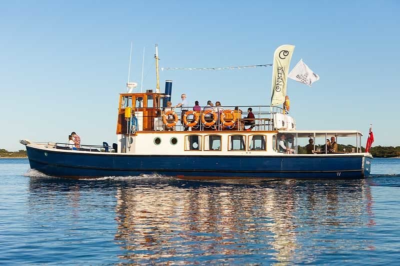 Old fashioned white and blue wooden steamer boat with a few people on the top deck and a Conker Spirit flag on the back in Poole Harbour on a sunny day. 